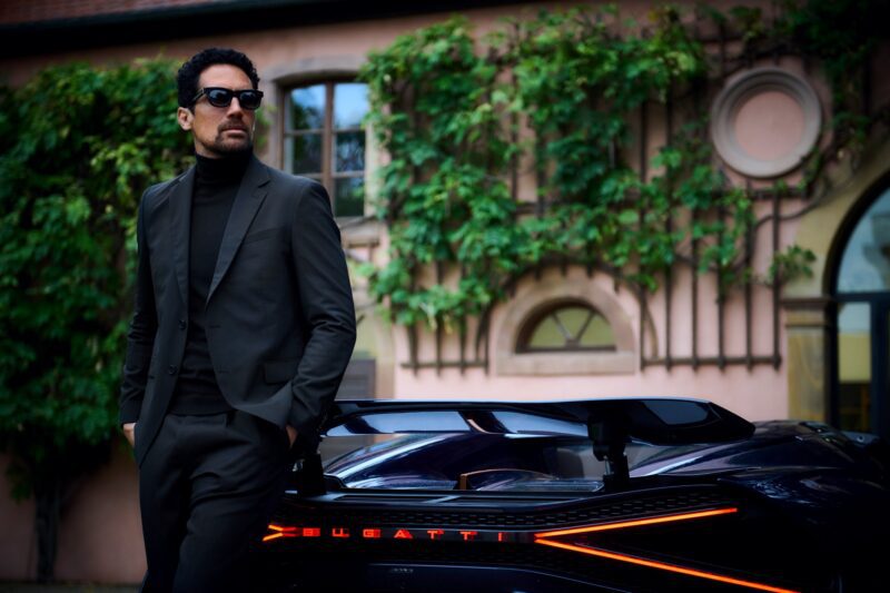 A man wearing a black suit and Bugatti eyewear stands next to a black Bugatti sports car in front of a building covered with climbing plants.