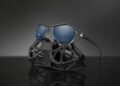 Bugatti Tourbillon-Inspired Eyewear with metallic frames and blue lenses rest on a geometric black display stand against a dark background, a striking preview for SILMO 2025.