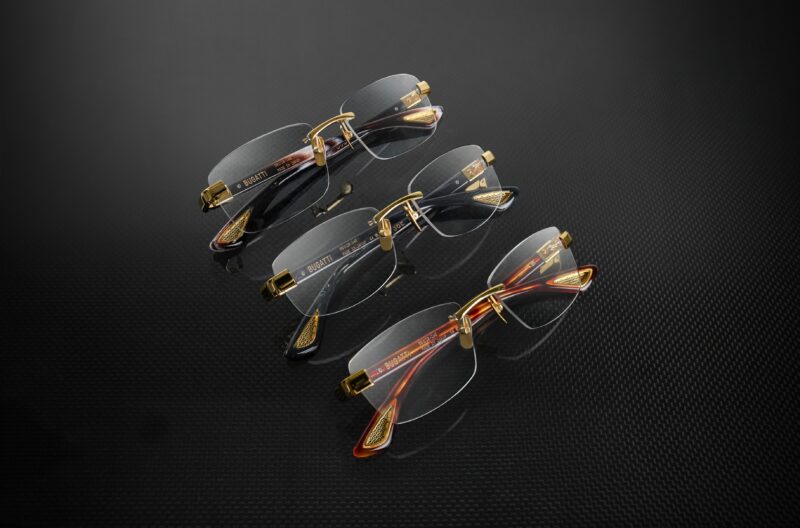 Three pairs of semi-rimless Bugatti eyeglasses with gold accents and transparent lenses are displayed on a dark, textured surface, showcasing Tourbillon-Inspired Eyewear elegance.
