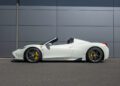An ultra-rare Ferrari 458 Speciale Aperta convertible with yellow brake calipers is parked in front of a gray metal-paneled wall on a paved surface.