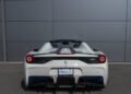 Rear view of a 2015 Ferrari 458 Speciale Aperta sports car parked in front of a gray metal wall, with dual exhaust pipes and a visible license plate holder. For sale.