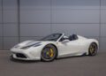 An ultra-rare Ferrari 458 Speciale Aperta, a white convertible sports car with yellow brake calipers, is parked on a paved surface in front of a gray paneled wall.