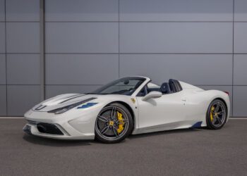 An ultra-rare Ferrari 458 Speciale Aperta, a white convertible sports car with yellow brake calipers, is parked on a paved surface in front of a gray paneled wall.