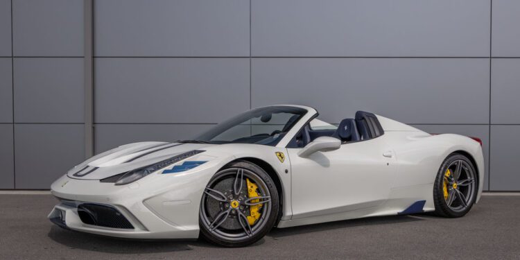 An ultra-rare Ferrari 458 Speciale Aperta, a white convertible sports car with yellow brake calipers, is parked on a paved surface in front of a gray paneled wall.