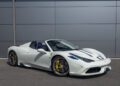 A 2015 Ferrari 458 Speciale Aperta convertible with blue racing stripes and yellow brake calipers is parked on a paved surface in front of a grey wall. Ferrari for sale.