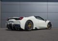 An ultra-rare Ferrari 458 Speciale Aperta with yellow brake calipers is parked on a clean asphalt surface in front of a gray, modern paneled wall.