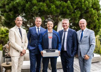 Five men in suits stand outdoors, posing for a photo. Abraham Cohen, at the center, holds a Bugatti Broward trophy for Best Performing Brand Manager. Green foliage and outdoor furniture are visible in the background.