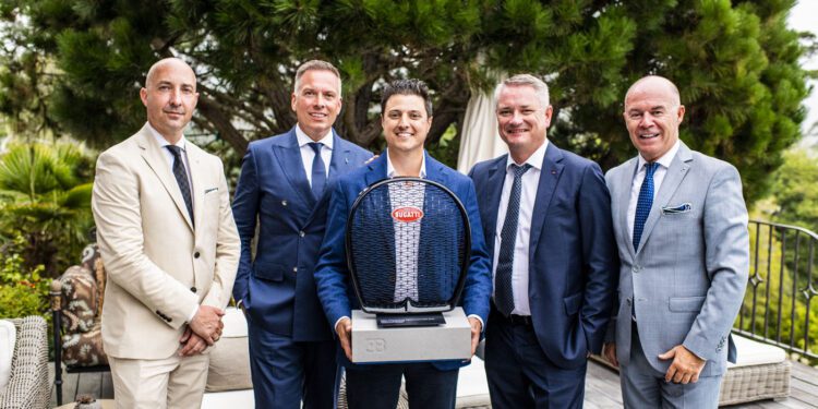 Five men in suits stand outdoors, posing for a photo. Abraham Cohen, at the center, holds a Bugatti Broward trophy for Best Performing Brand Manager. Green foliage and outdoor furniture are visible in the background.