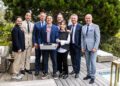 A group of eight people, including Abraham Cohen, Best Performing Brand Manager at Bugatti Broward, dressed in business and semi-formal attire, pose together on a wooden deck outdoors; one person holds a tennis racket and a box.