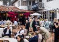 A crowd of people socialize at an outdoor gathering on a tiled patio with comfortable seating, flowers, and a "Bugatti Broward" sign in the background.