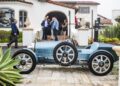A vintage blue car from Bugatti Broward is parked on a stone driveway in front of a large house, with several people gathered and talking in the background—including Best Performing Brand Manager Abraham Cohen.