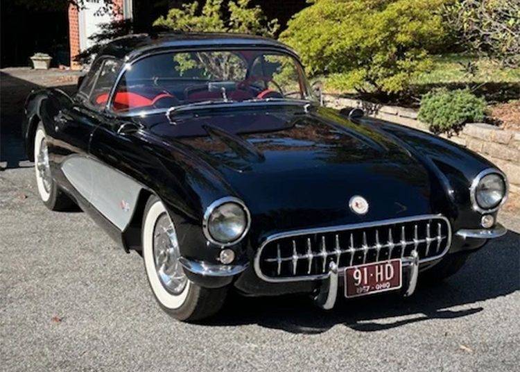 A low mileage classic Chevrolet Corvette convertible with black paint, white side panels, and red interior is parked on a driveway near greenery.
