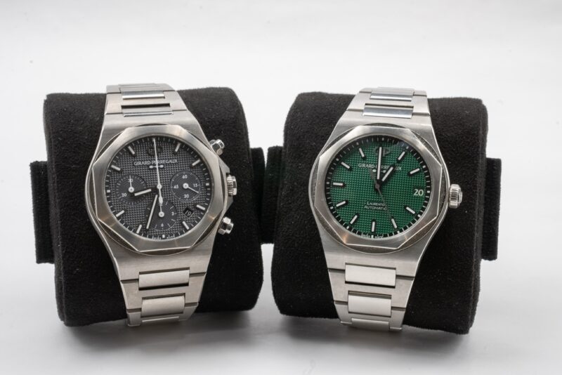 Two stainless steel wristwatches on black cushions; the left watch is a Girard-Perregaux with a black chronograph dial, while the right boasts a green textured dial and date display—a duo perfect for Monterey Car Week and Time Well Spent.