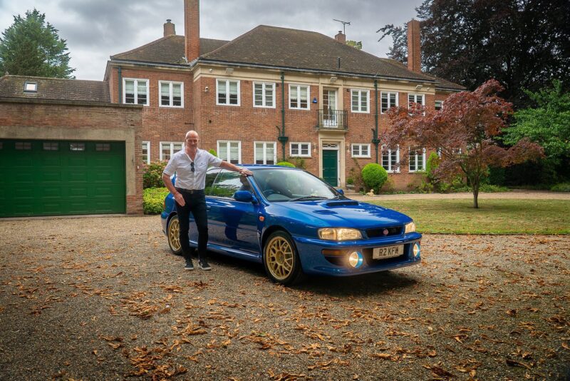 A person stands next to a blue Subaru 22B STi with gold wheels parked in front of a large brick house with green garage doors, perfect for a Petrolicious Film Friday feature.