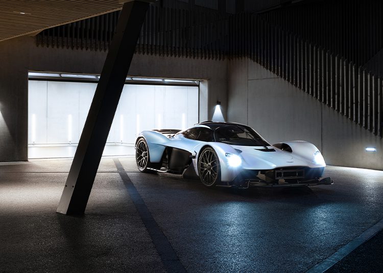 A silver, low-profile Aston Martin Valkyrie with headlights on is parked in a modern, dimly lit garage with concrete walls and geometric elements—an auction-worthy scene reminiscent of Daniel Ricciardo’s racing flair.