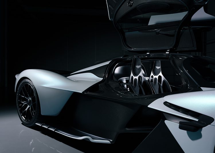 A sleek, white and black Aston Martin Valkyrie sports car with gullwing door open, revealing two racing seats inside, set against a dark background—perfect for auction or fans of Daniel Ricciardo.
