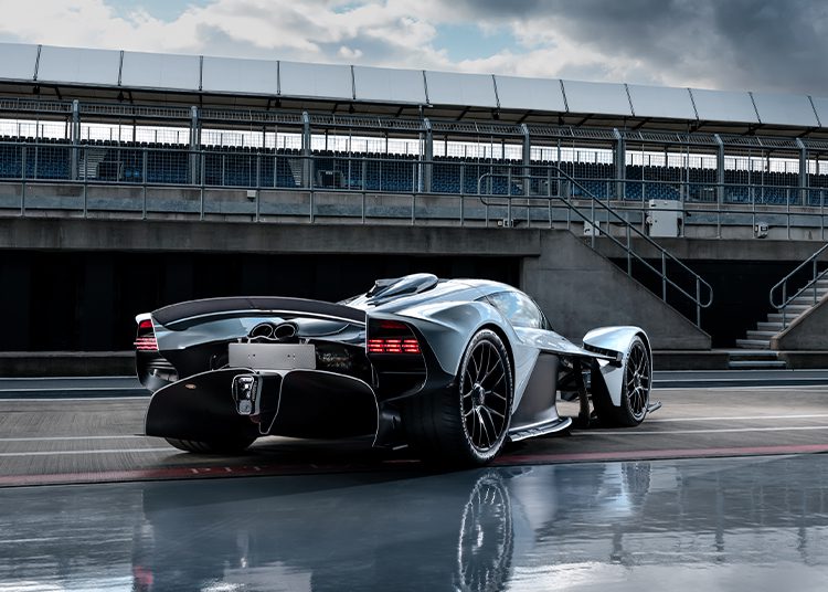 A sleek silver Aston Martin Valkyrie hypercar is parked on a wet racetrack in front of empty grandstands under a cloudy sky.