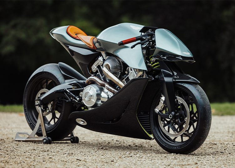 A modern, custom-built motorcycle with a sleek, aerodynamic design and an orange seat, displayed on a stand outdoors—perfect for fans of auction excitement or Daniel Ricciardo’s passion for engineering excellence.