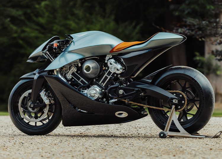 A modern, futuristic motorcycle with a matte silver body, exposed engine components, and a single rear stand, parked on a gravel surface with greenery in the background—reminiscent of the innovation seen in the Aston Martin Valkyrie.