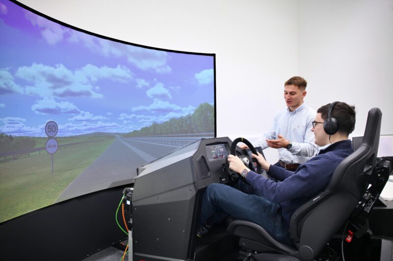 In a tech center, a person wearing headphones operates a driving simulator with a curved screen, while another explains nearby—demonstrating cutting-edge Bentley electrification technology.