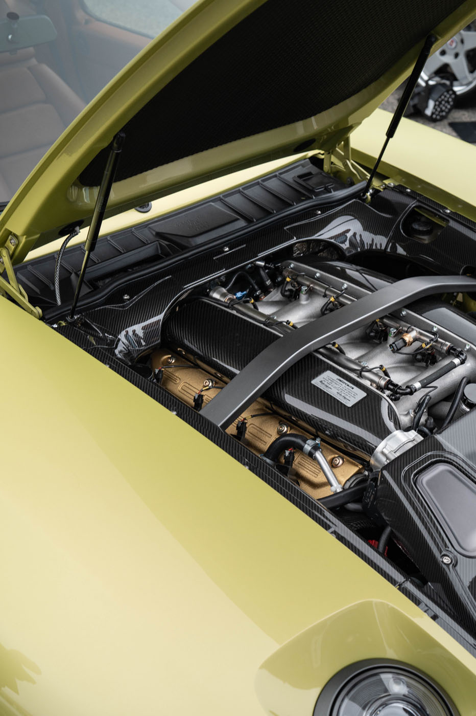 A close-up view of a car engine with carbon fiber and gold components under the hood of a yellow Nardone 928 production car.