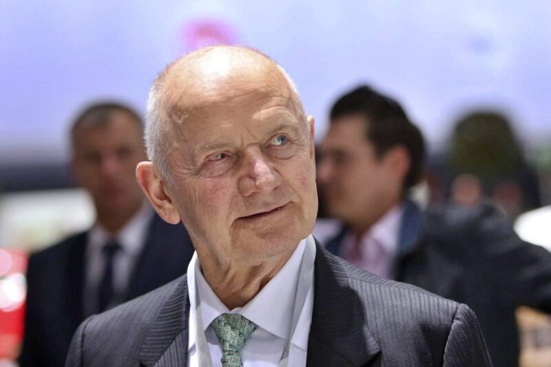 An elderly man in a suit and tie looks upward, with blurred people in business attire in the background—much like how Bugatti honors the Veyron’s 20-year legacy with a special logo, blending tradition and innovation.