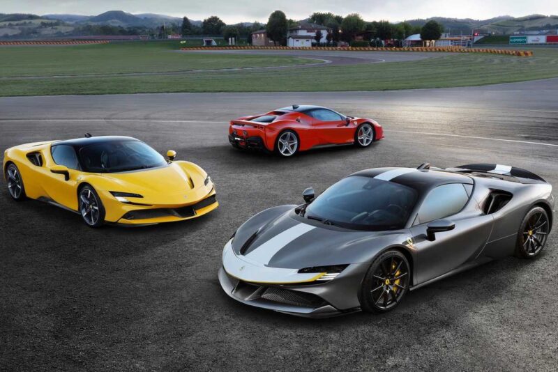 Three sports cars—one yellow, one red, and one gray with black stripes—are parked on an asphalt surface near a racetrack, setting the scene for an exciting comparison: Ferrari SF90 Stradale vs. Ferrari 849 Testarossa.