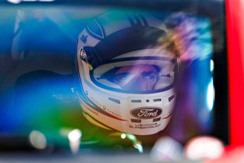 Close-up of a race car driver wearing a white Ford helmet, seen through a car window with colorful reflections—a nod to the spirit of Two Fords EVs Two Records: Lightning SuperTruck and SuperVan conquer the Green Hell.
