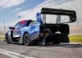 A blue and black electric race car, inspired by the Lightning SuperTruck and SuperVan that conquered the Green Hell, is parked on a race track near a fence, its large rear wing and exposed rear components on full display.