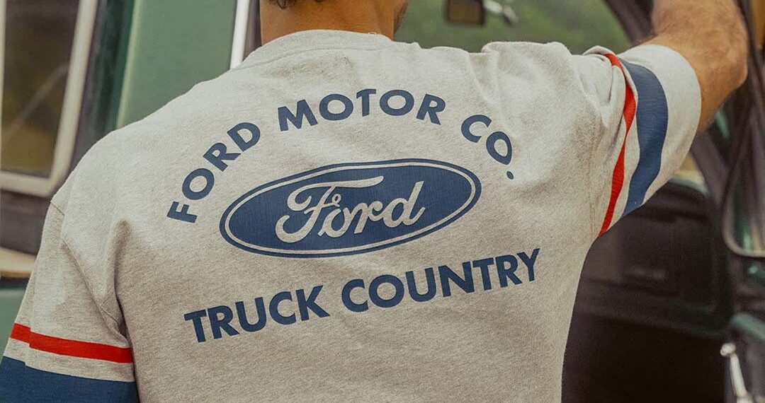 A person wearing a "Ford x Brixton Limited Edition Collection" t-shirt stands beside an open green vehicle door, facing away from the camera.