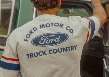 A person wearing a "Ford x Brixton Limited Edition Collection" t-shirt stands beside an open green vehicle door, facing away from the camera.