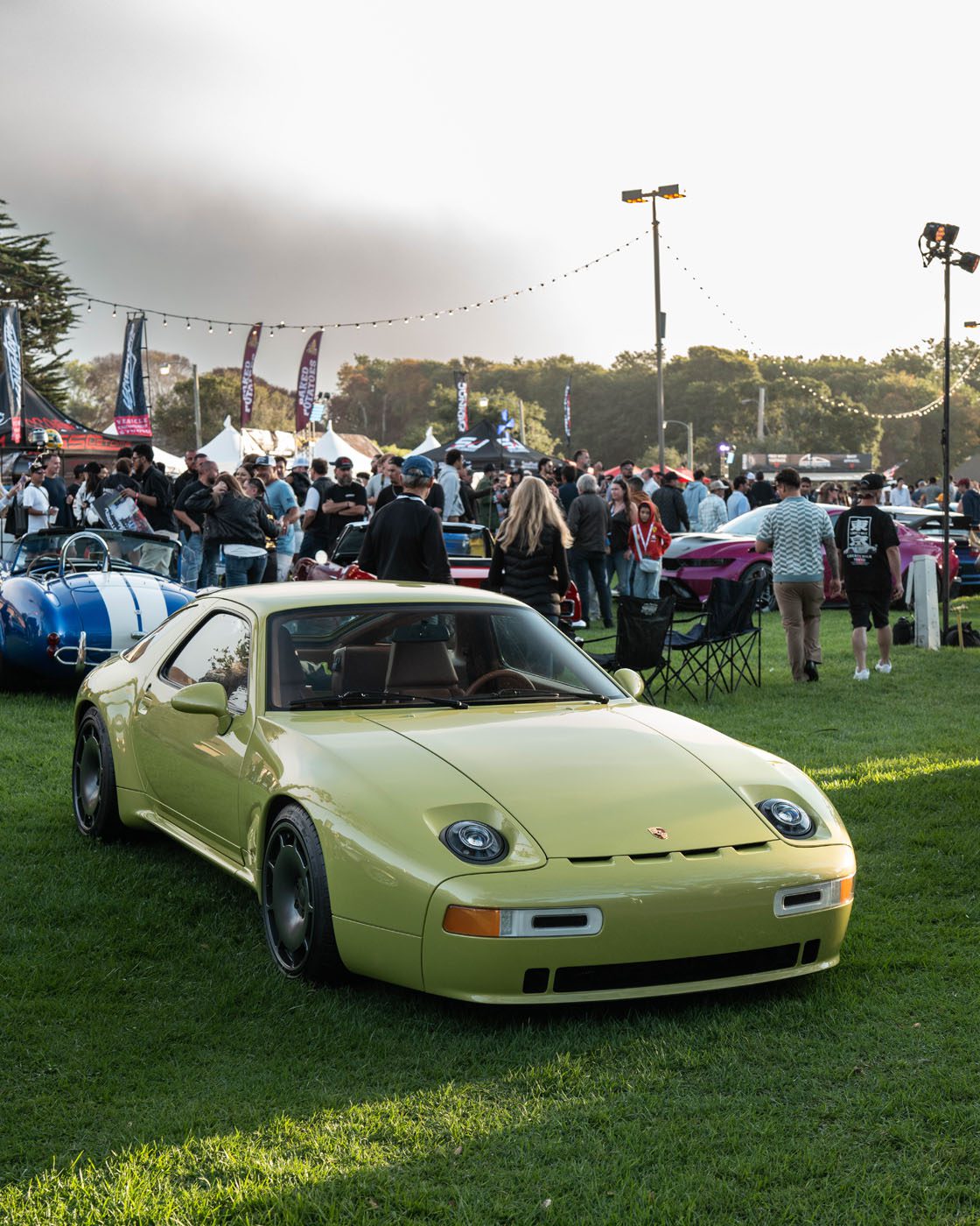 A yellow Nardone 928 is parked on grass at a car show, with people gathered and other GH Vehicles production cars displayed in the background.