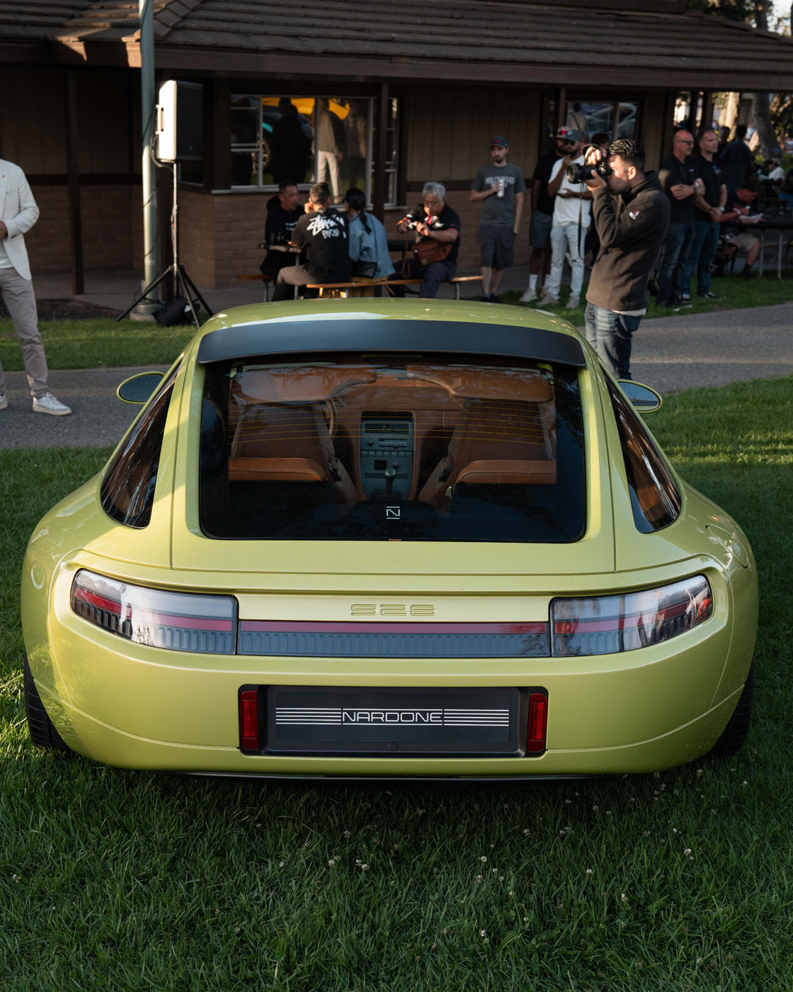 A green Nardone 928 modern sports car is displayed on grass with people standing and sitting in the background, some taking photos and filming.