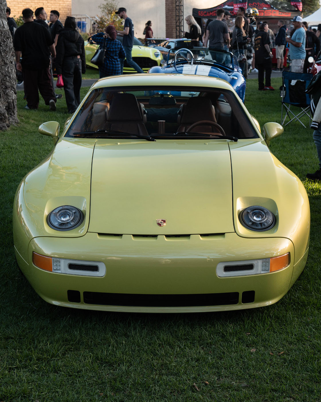 A yellow Nardone 928 is parked on grass at an outdoor car show, with people and other production cars visible in the background.