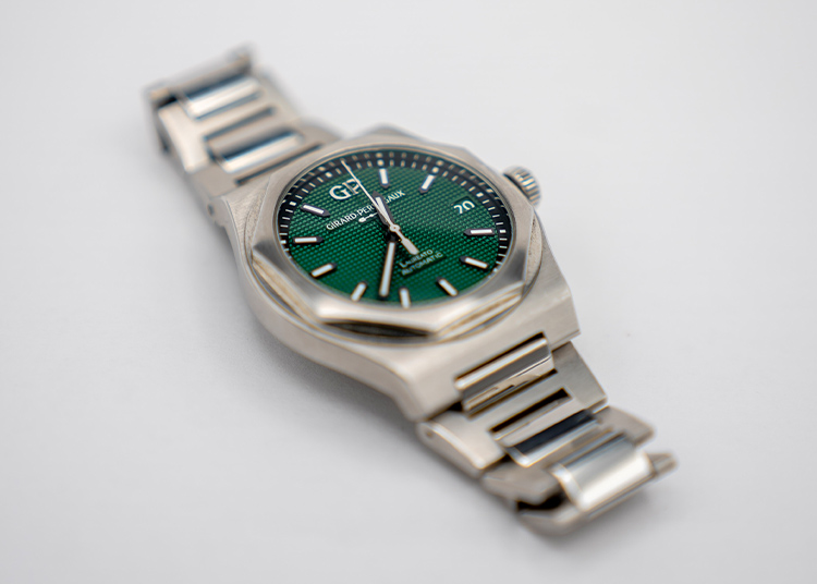 A silver wristwatch with a green textured dial, silver hour markers, and a metal link band evokes the elegance of Girard-Perregaux, making every moment Time Well Spent against a plain white background.