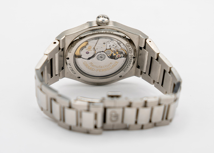 A silver Girard-Perregaux wristwatch with an open case back displays its intricate mechanical movement and engraved branding on a white background—a true symbol of Time Well Spent.
