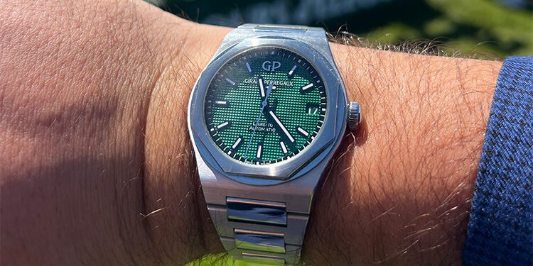 A Girard-Perregaux stainless steel wristwatch with a green textured dial and silver hour markers is worn on a person's wrist outdoors—perfect for making Monterey Car Week memories, reminding you that it's always time well spent.