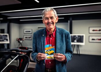 An older man in a blue blazer and red shirt holds a yellow Hot Wheels T.33 package in a room with framed photos, cars, and a nod to Gordon Murray in the background.