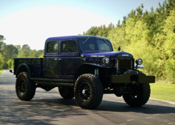 A restored 1947 Desert Power Wagon with large off-road tires is parked on a paved road, surrounded by greenery and trees—a striking tribute to classic trucks enhanced with Turbo Diesel power.
