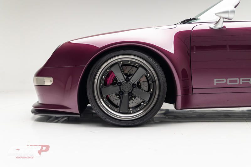 Side view of a maroon sports car's front left wheel and fender, featuring black alloy rims and visible brake calipers, inspired by the sleek design of the Gunther Werks Speedster, against a white background.