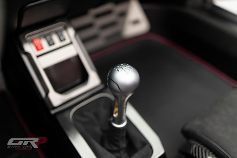 Close-up of a manual car gear shift knob with visible gear pattern, surrounded by a black and silver console in a bespoke Gunther Werks Speedster, enhanced by Graham Rahal Performance touches.
