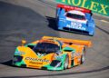 Two vintage race cars—a green-orange Mazda and a blue Nissan—celebrate Japanese Motorsports Heritage as they speed past a green Rolex ad at the 2026 Rolex Monterey Motorsports Reunion.