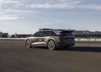 A black SUV with logos including Lucid, Nuro, and Uber drives on a highway, with city buildings and mountains in the background.
