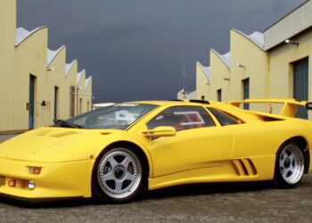 A yellow Lamborghini Diablo—celebrating 35 years as the last old-school analog Raging Bull—stands parked between yellow industrial buildings with blue doors, its legendary roar echoing under a cloudy sky.
