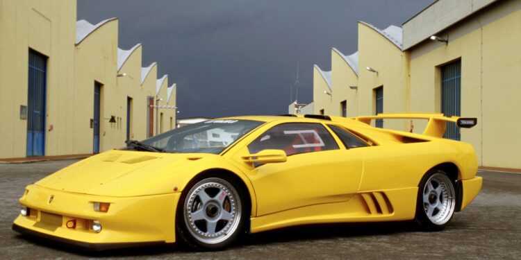 A yellow Lamborghini Diablo—celebrating 35 years as the last old-school analog Raging Bull—stands parked between yellow industrial buildings with blue doors, its legendary roar echoing under a cloudy sky.