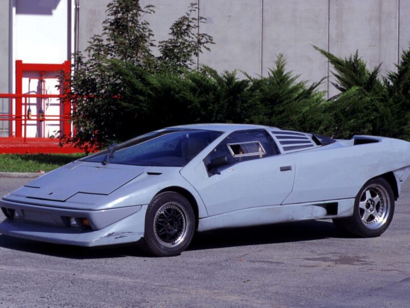 A silver wedge-shaped Lamborghini Diablo turns 35, parked outdoors on concrete near green bushes and a gray building—the last old-school analog Raging Bull still roars.
