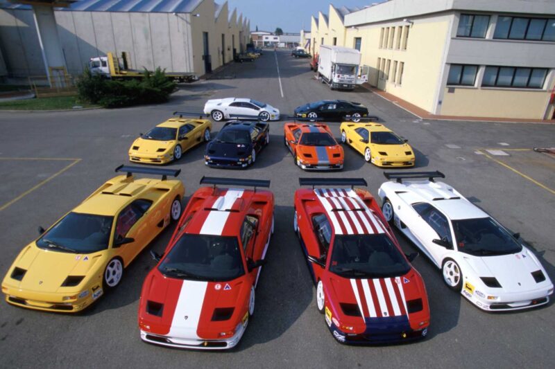 A group of ten Lamborghini sports cars, mostly Diablo models, are parked in a lot between industrial buildings—proof the Lamborghini Diablo turns 35: the last old-school analog raging bull still roars.