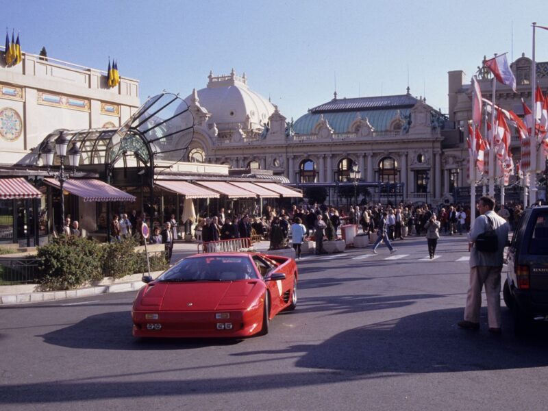 A red Lamborghini Diablo, celebrating 35 years as the last old-school analog Raging Bull, drives past a crowd outside an ornate building with domes and striped awnings on a sunny day.