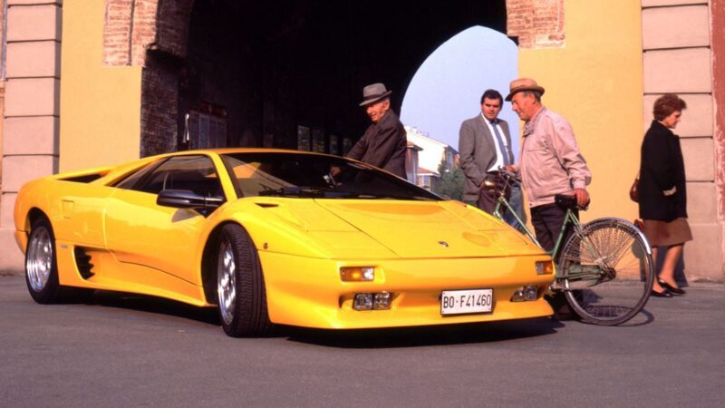 A yellow Lamborghini Diablo, the last old-school analog Raging Bull, is parked on the street near an archway as three men and a woman stand nearby, one holding a bicycle. Lamborghini Diablo Turns 35, and it still roars with timeless style.
