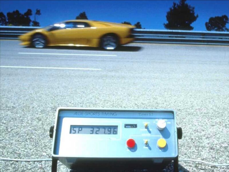 A yellow Lamborghini Diablo speeds past a guardrail, its timing device displaying numbers in the foreground as the last old-school analog raging bull still roars on a sunny day.